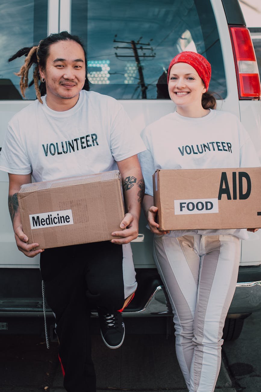 man and woman carrying medicine and food labelled cardboard boxes behind a white van